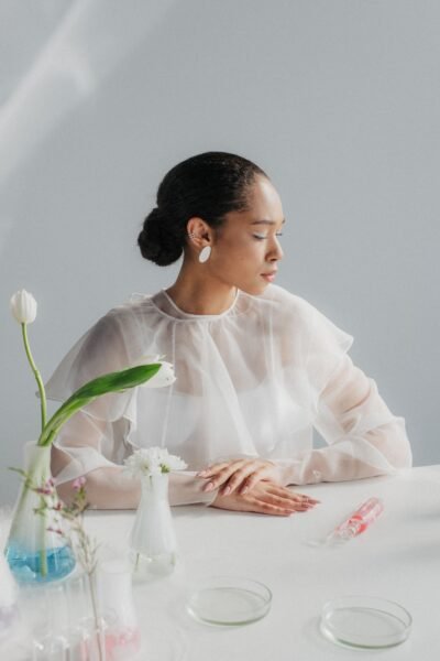 Woman in a sheer blouse sitting in a minimalist laboratory setting with glassware.