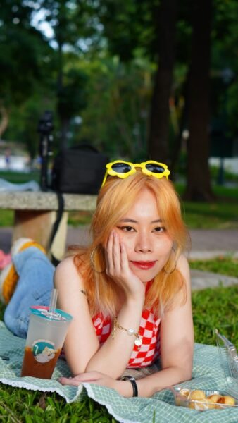 Woman with blonde hair and sunglasses relaxing on a picnic blanket with a drink in a park.