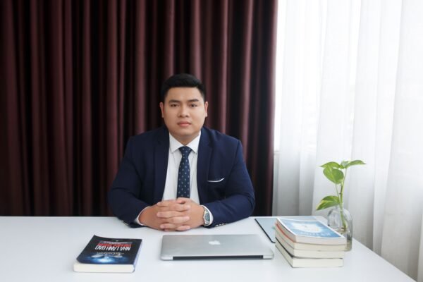 Businessman in a formal suit sitting at a desk with a laptop and books in a modern office setting.