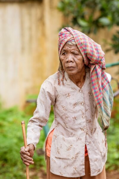 A senior woman wearing traditional attire and headscarf, holding a stick, in Thị xã Phước Long, Bình Phước, Vietnam.