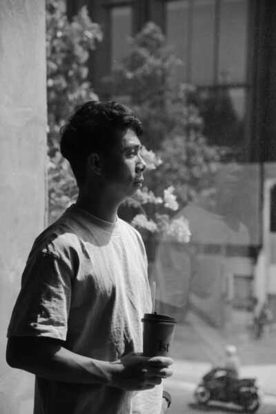 Black and white portrait of a young man holding a coffee cup and looking out of a cafe window.